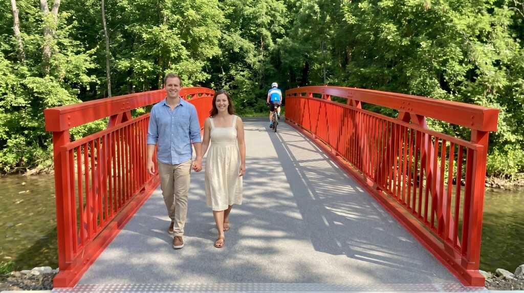 Ein Pärchen spaziert auf einer roten Aluminiumbrücke und lächelt in die Kamera