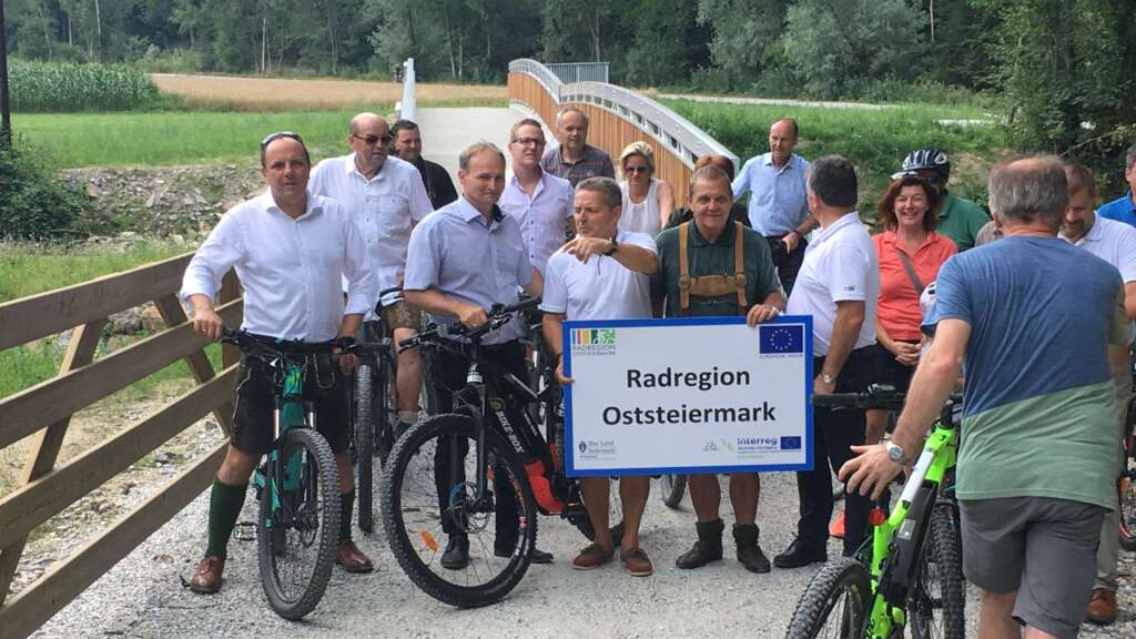 Politiker mit dem Schild „Radregion Oststeiermark“ posieren vor zwei neu errichteten 18,50 m langen Aluminiumbrücken am Radweg R8 und freuen sich über die Eröffnung des neuen Radwegabschnitts.