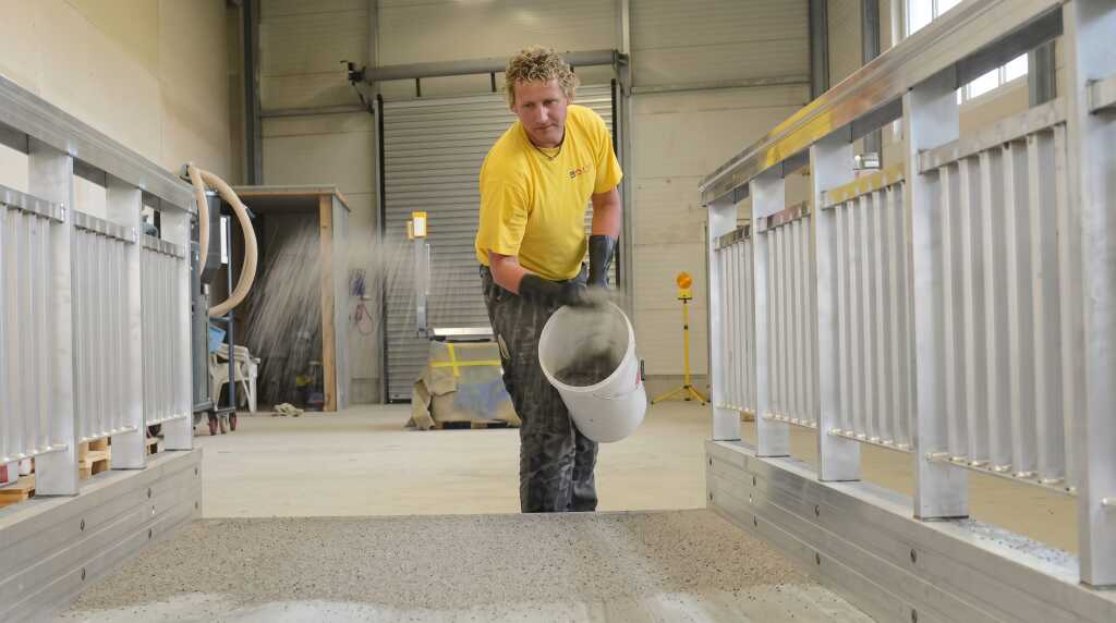 Mitarbeiter streut Sand in eine frisch aufgetragene Polyurethanbeschichtung auf einer Aluminiumbrücke.